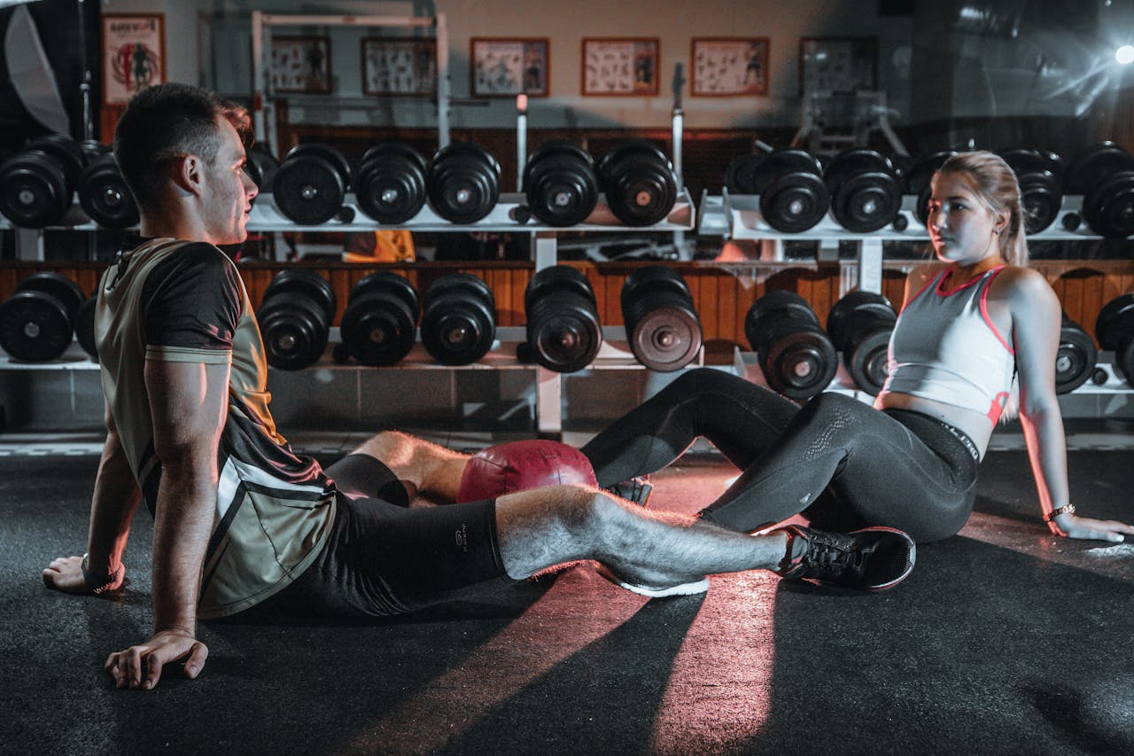 hero-img-02 A man and woman resting together on the gym floor, surrounded by dumbbells.